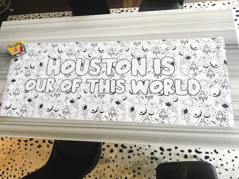 Może przedstawiać: Czarno-biała kolorowanka z motywem kosmicznym, przedstawiająca rakiety, planety i astronaut&oacute;w. Tekst "HOUSTON IS OUT OF THIS WORLD" jest wyraźnie widoczny. W rogu widoczne jest pudełko kredek.