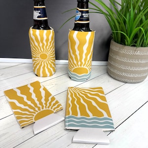May include: Yellow and white sunburst design beer can coolers and matching can holders. The design features a yellow sun with white rays. The bottom of the design has a blue wave pattern. Two beer bottles are in the coolers.