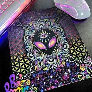 May include: A colorful mousepad featuring a black alien head with a third eye, surrounded by a mandala design. The background has a galaxy and geometric pattern. The mousepad is next to a white keyboard and mouse.