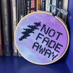 May include: A cross-stitch embroidery hoop with the words "NOT FADE AWAY" in black thread. The background is a gradient of purple and blue. A black tree-like design is above the text. The hoop is clamped to a shelf.