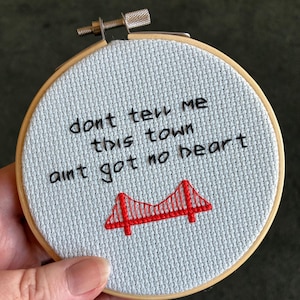 May include: A cross-stitch embroidery hoop with the text "don't tell me this town ain't got no heart" and a red bridge design. The text and bridge are stitched in black and red thread on a light blue fabric, set in a wooden hoop.