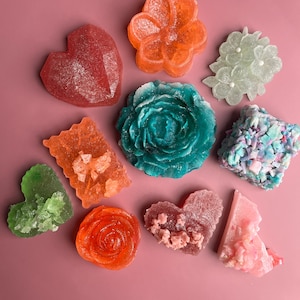 Puede incluir: Un surtido de jabones decorativos coloridos en varias formas, incluyendo corazones, flores y una rosa. Los jabones son en tonos de rojo, naranja, verde, azul y rosa, dispuestos sobre una superficie rosa. Algunos jabones tienen una textura cristalina.