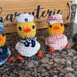 May include: Four handmade crochet ducklings with yellow bodies, orange beaks and feet, and black eyes. Each duckling wears a different outfit: a sailor hat and scarf, a blue cap and sweater, a pink and white striped outfit, and a green hat and sweater.