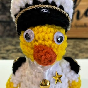 May include: A crocheted yellow duck wearing a white and black sailor hat with a gold trim and a gold star. The duck is wearing a white crocheted uniform with gold buttons and an orange crocheted collar.