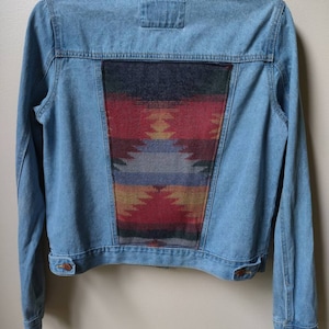 Puede incluir: Una chaqueta vaquera azul claro con un parche de tela estampado de estilo suroeste en la parte posterior.
