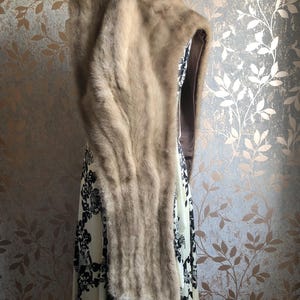 May include: A brown fur hooded scarf with a long tassel. The scarf is draped over a white dress with black floral print.