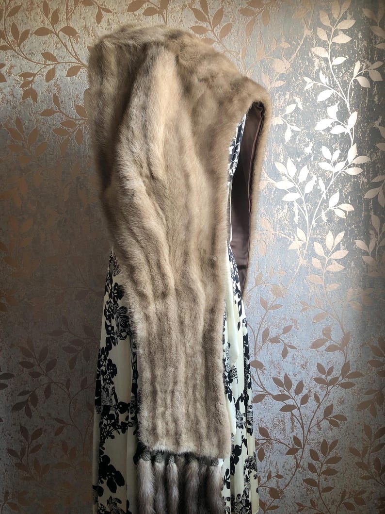 May include: A brown fur hooded scarf with a long tassel. The scarf is draped over a white dress with black floral print.