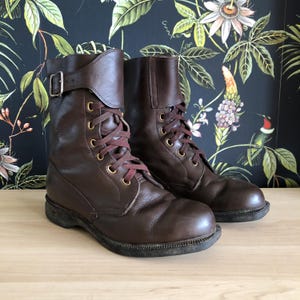 Könnte beinhalten: Ein Paar dunkelbraune Lederstiefel in wadenhoher Ausführung mit abgerundeten Zehenkappen und einer Schnürung mit messingfarbenen Ösen. Jeder Stiefel verfügt über einen verstellbaren Riemen am oberen Schaft, der mit einer silberfarbenen Metallschnalle schließt.