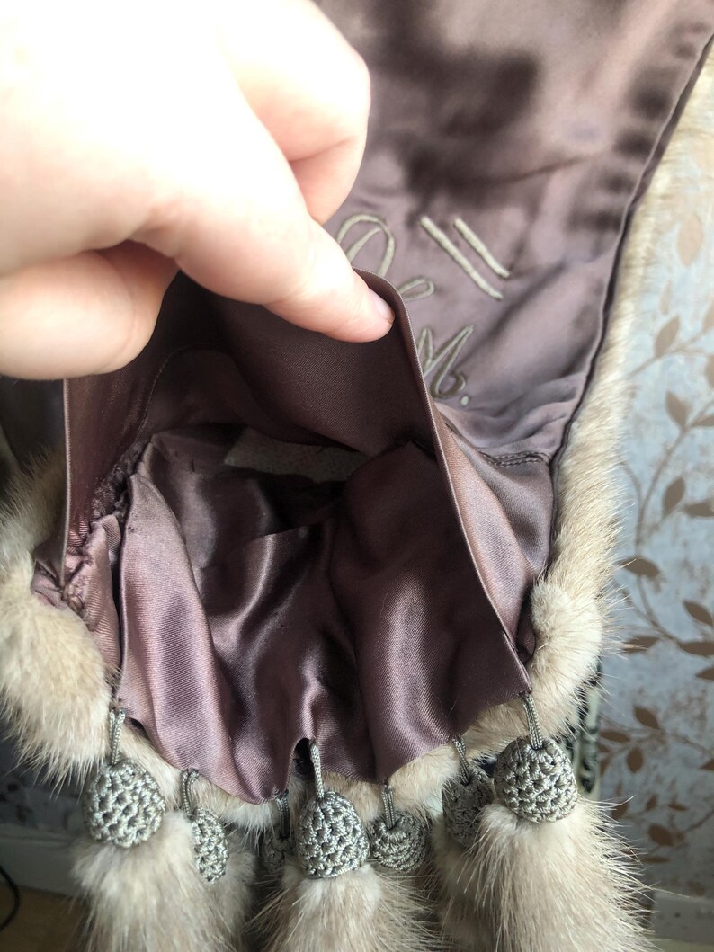 May include: A brown fur stole with a satin lining and a grey crocheted tassel trim. The lining has the text "21 M." embroidered on it.