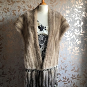 May include: A light brown fur shawl with a fringe of fur tails. The shawl is displayed on a white mannequin against a patterned background.