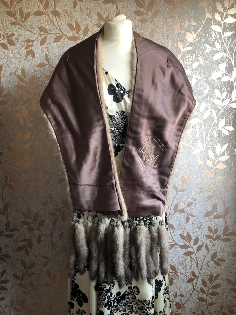 May include: A brown fur stole with a satin lining and a fringe of fur tails. The stole is embroidered with the initials "D.M."