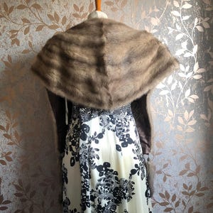 May include: A brown fur shawl with a long, flowing design. The shawl is draped over a white dress with a black floral pattern.