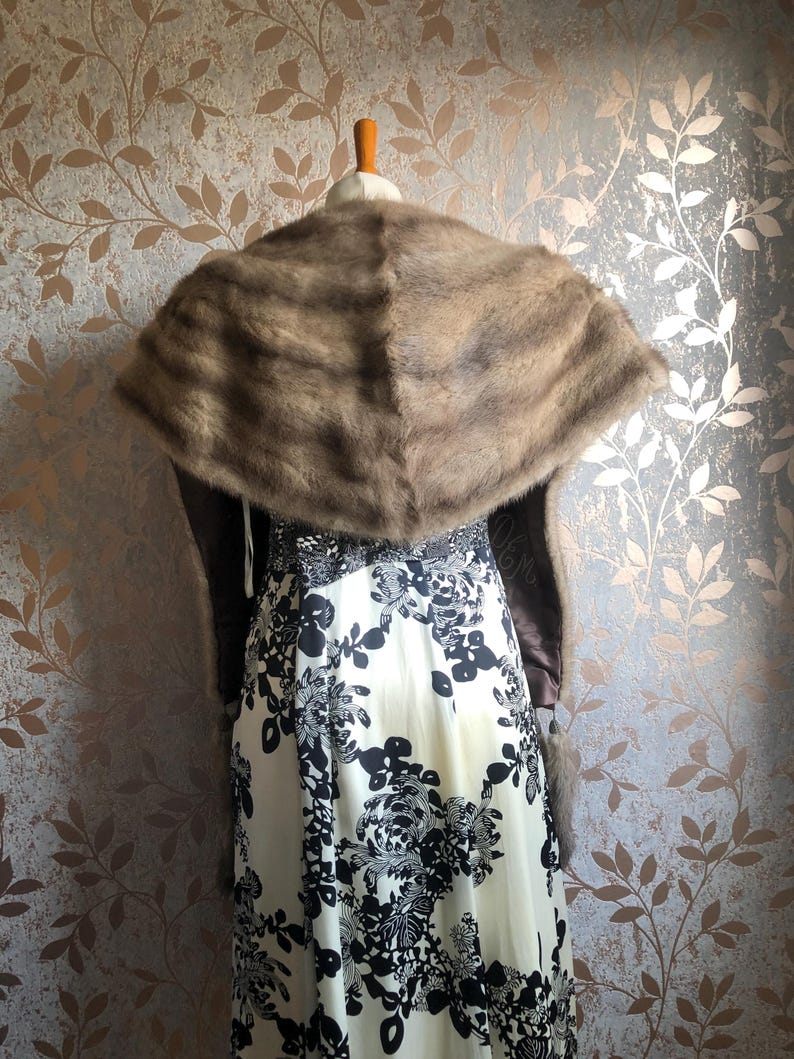 May include: A brown fur shawl with a long, flowing design. The shawl is draped over a white dress with a black floral pattern.