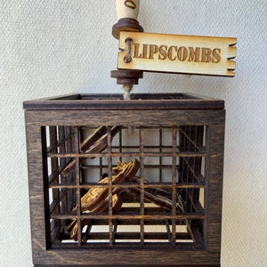 May include: A wooden cage ornament with a dark brown finish, featuring a rectangular shape with a grid design. The ornament includes a wooden tag that reads "LIPSCOMBS" and a wooden handle with the year "2020" engraved on it.