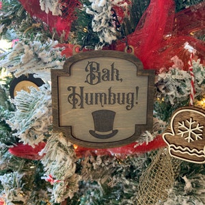 May include: Wooden Christmas tree ornament with the phrase "Bah, Humbug!" and a top hat illustration. The ornament is brown and gray, hanging on a Christmas tree with red ribbon and faux snow.