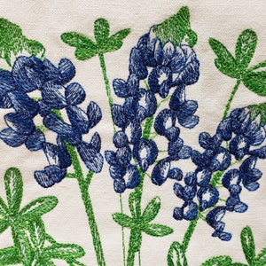 May include: Embroidered bluebonnet flowers with green leaves on a white fabric background.