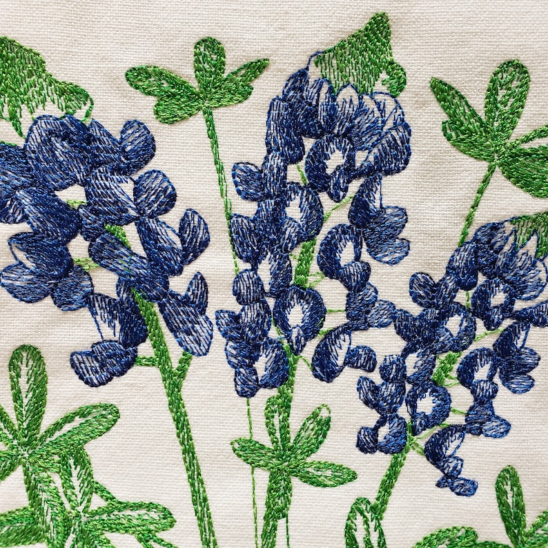 May include: Embroidered bluebonnet flowers with green leaves on a white fabric background.