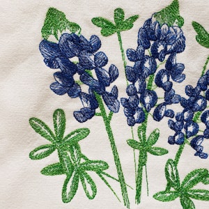 May include: Embroidered bluebonnet flowers with green leaves on a white fabric background.