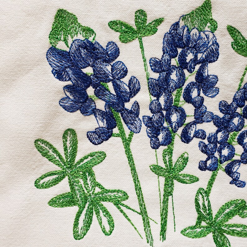 May include: Embroidered bluebonnet flowers with green leaves on a white fabric background.