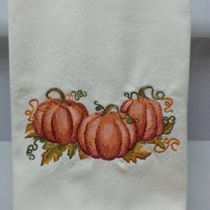 May include: Off-white kitchen towel with an embroidered design of three orange pumpkins. The pumpkins have green stems and are surrounded by fall leaves and orange vines. The towel is made of a soft, absorbent material.