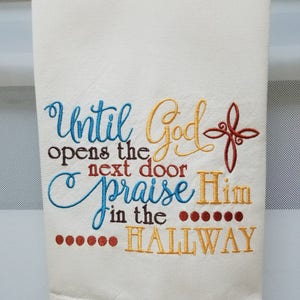 May include: White kitchen towel with embroidered text in blue, yellow, orange, and brown. The text reads "Until God opens the next door praise Him in the hallway".
