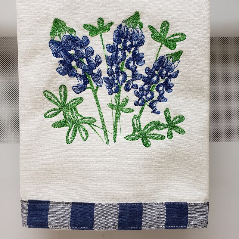 May include: A white kitchen towel with a blue and white checkered border. The towel features an embroidered design of blue and green Texas bluebonnets.
