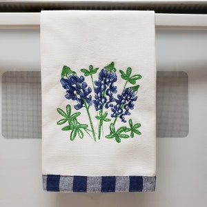May include: A white kitchen towel with a blue and white checkered border. The towel features an embroidered design of bluebonnet flowers in shades of blue and green.