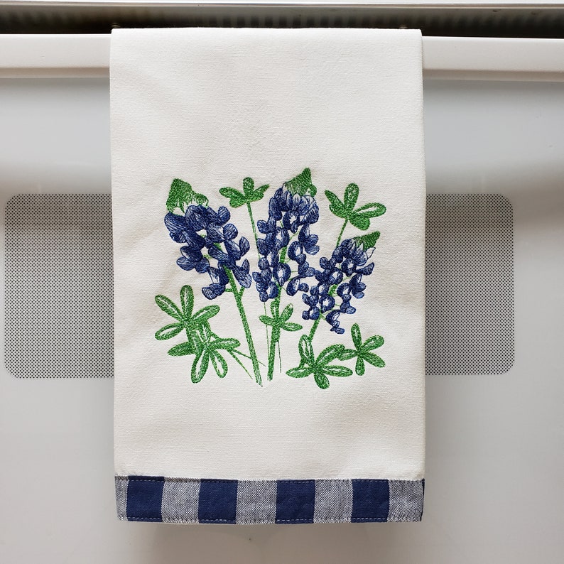 May include: A white kitchen towel with a blue and white checkered border. The towel features an embroidered design of bluebonnet flowers in shades of blue and green.