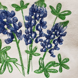 May include: Embroidered design of bluebonnet flowers with green leaves on a white fabric background.