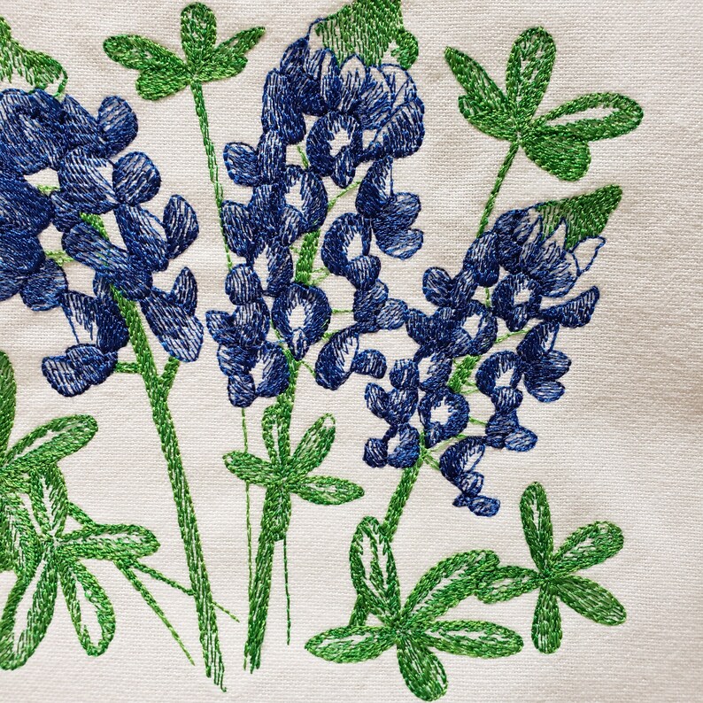 May include: Embroidered design of bluebonnet flowers with green leaves on a white fabric background.