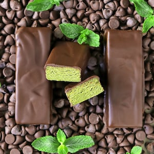 May include: Three chocolate bars with a green mint filling are arranged on a bed of chocolate chips. The bars are covered in dark chocolate and have a smooth, glossy finish. Fresh mint leaves are scattered around the bars.