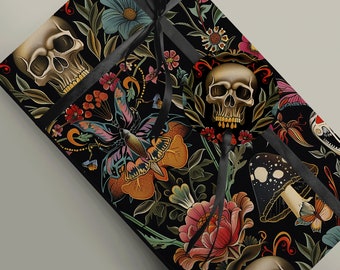 May include: A rectangular gift box wrapped in black paper featuring a colourful pattern of skulls, butterflies, mushrooms, and flowers. A black satin ribbon is tied around the box, securing a circular gift tag with a skull illustration on the front.