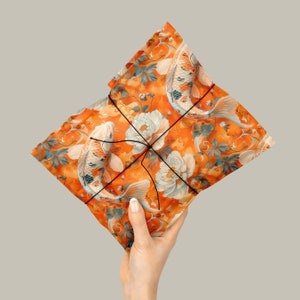 Puede incluir: Papel de regalo naranja con un patr&oacute;n de peces koi y peon&iacute;as blancas. El papel est&aacute; atado con una cuerda negra.