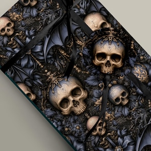 May include: A rectangular gift box wrapped in paper featuring a repeating pattern of beige human skulls, dark blue bat wings, and intricate floral elements. A black satin ribbon is tied around the exterior of the box, crossing over the surface.