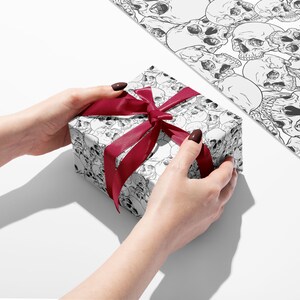 Wrapping paper featuring skulls showing hands holding a gift with red ribbon and a flat sheet of wrapping paper.