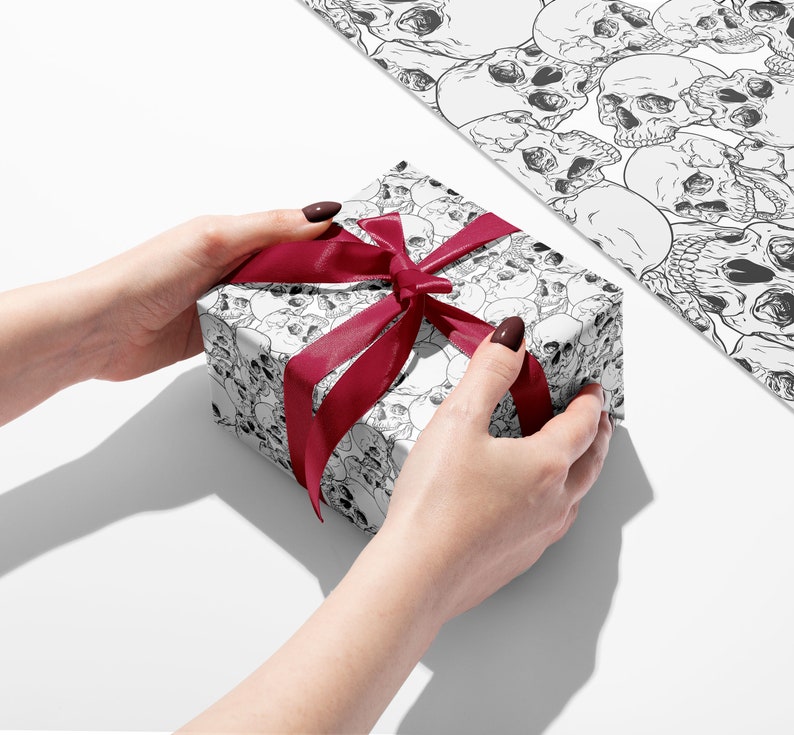 Wrapping paper featuring skulls showing hands holding a gift with red ribbon and a flat sheet of wrapping paper.