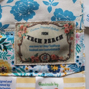 May include: A close-up of a fabric label with the text "Custom Printed Label" and "FROM TECH PERCH" on a white background with floral designs. The label is attached to a fabric bag with a blue and white floral pattern.