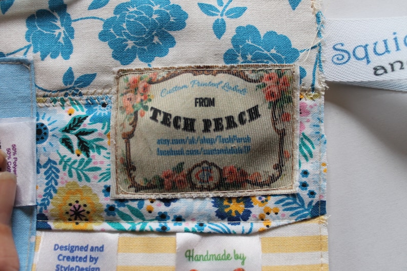May include: A close-up of a fabric label with the text "Custom Printed Label" and "FROM TECH PERCH" on a white background with floral designs. The label is attached to a fabric bag with a blue and white floral pattern.