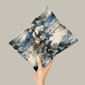 May include: A rectangular package wrapped in paper featuring a detailed landscape illustration with blue mountains, white flowering trees, and traditional buildings. The paper is secured with thin black string tied in a cross pattern.