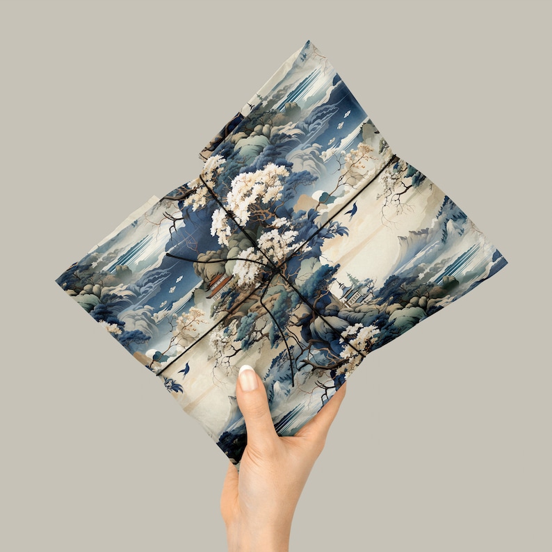 May include: A rectangular package wrapped in paper featuring a detailed landscape illustration with blue mountains, white flowering trees, and traditional buildings. The paper is secured with thin black string tied in a cross pattern.