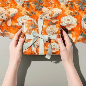 Puede incluir: Una caja de regalo envuelta en papel estampado naranja y blanco con peces koi y flores. La caja est&aacute; atada con una cinta verde claro.