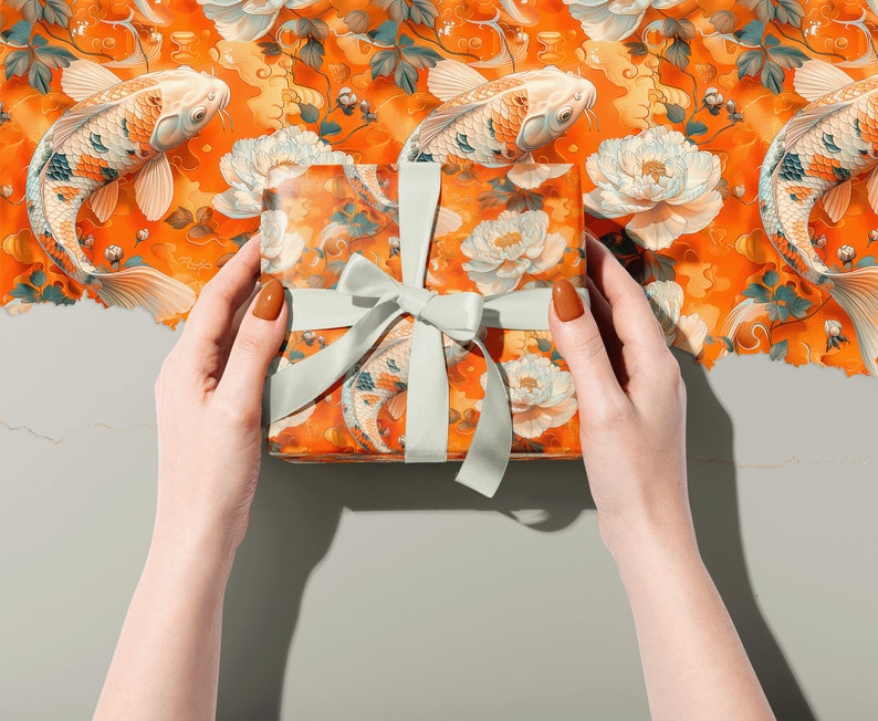 Puede incluir: Una caja de regalo envuelta en papel estampado naranja y blanco con peces koi y flores. La caja est&aacute; atada con una cinta verde claro.