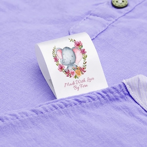 May include: A white fabric label with a cute cartoon elephant surrounded by pink and yellow flowers. The label says "Made With Love By Fern".