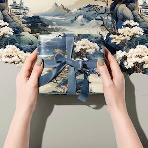 May include: A square gift box wrapped in paper featuring a detailed landscape illustration of mountains, trees, and pagodas in shades of blue, cream, and white, tied with a solid slate blue satin ribbon bow centred on the top surface.