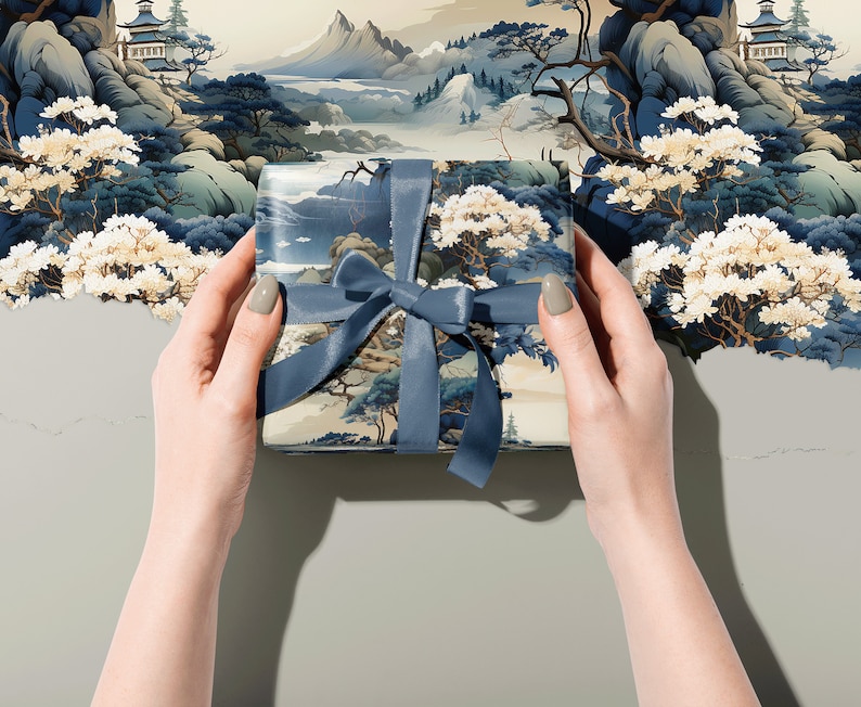May include: A square gift box wrapped in paper featuring a detailed landscape illustration of mountains, trees, and pagodas in shades of blue, cream, and white, tied with a solid slate blue satin ribbon bow centred on the top surface.