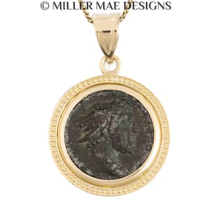 May include: A gold-colored pendant necklace featuring a circular coin with a detailed profile image. The coin is set within a gold bezel with a beaded border. The necklace is suspended from a delicate gold chain. The text "© MILLER MAE DESIGNS" is at the top.