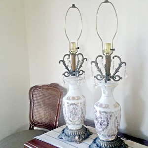 May include: Pair of white ceramic table lamps with floral designs and ornate metal accents. Each lamp features a decorative metal filigree, crystal-like dangles, and a brass-colored base. The lamps are approximately 24 inches tall.