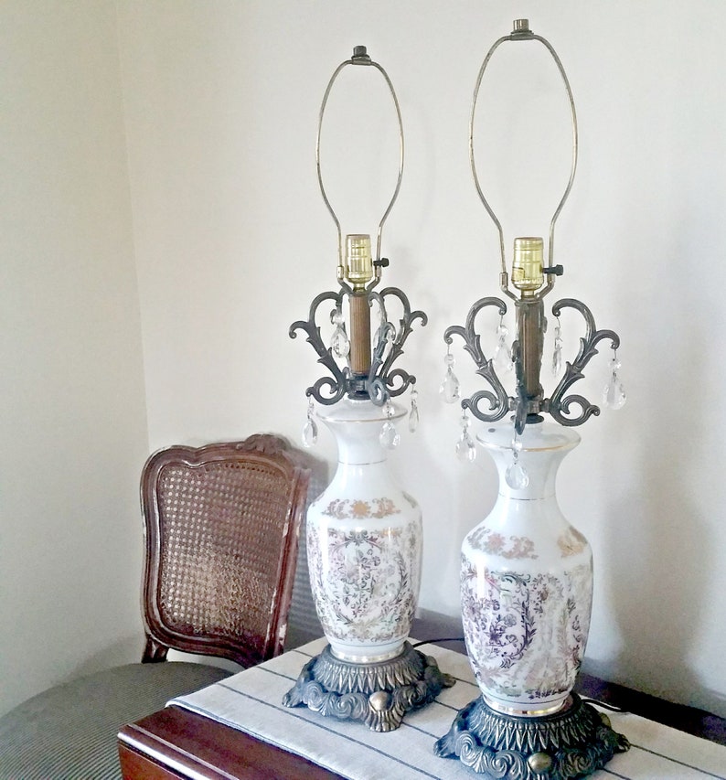May include: Pair of white ceramic table lamps with floral designs and ornate metal accents. Each lamp features a decorative metal filigree, crystal-like dangles, and a brass-colored base. The lamps are approximately 24 inches tall.