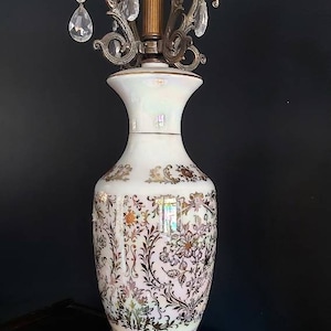 May include: An ornate table lamp with a white ceramic base featuring a floral pattern in gold and brown. The lamp has a brass-colored metal base and a decorative top with hanging crystal accents. The lamp's design is vintage and elegant.