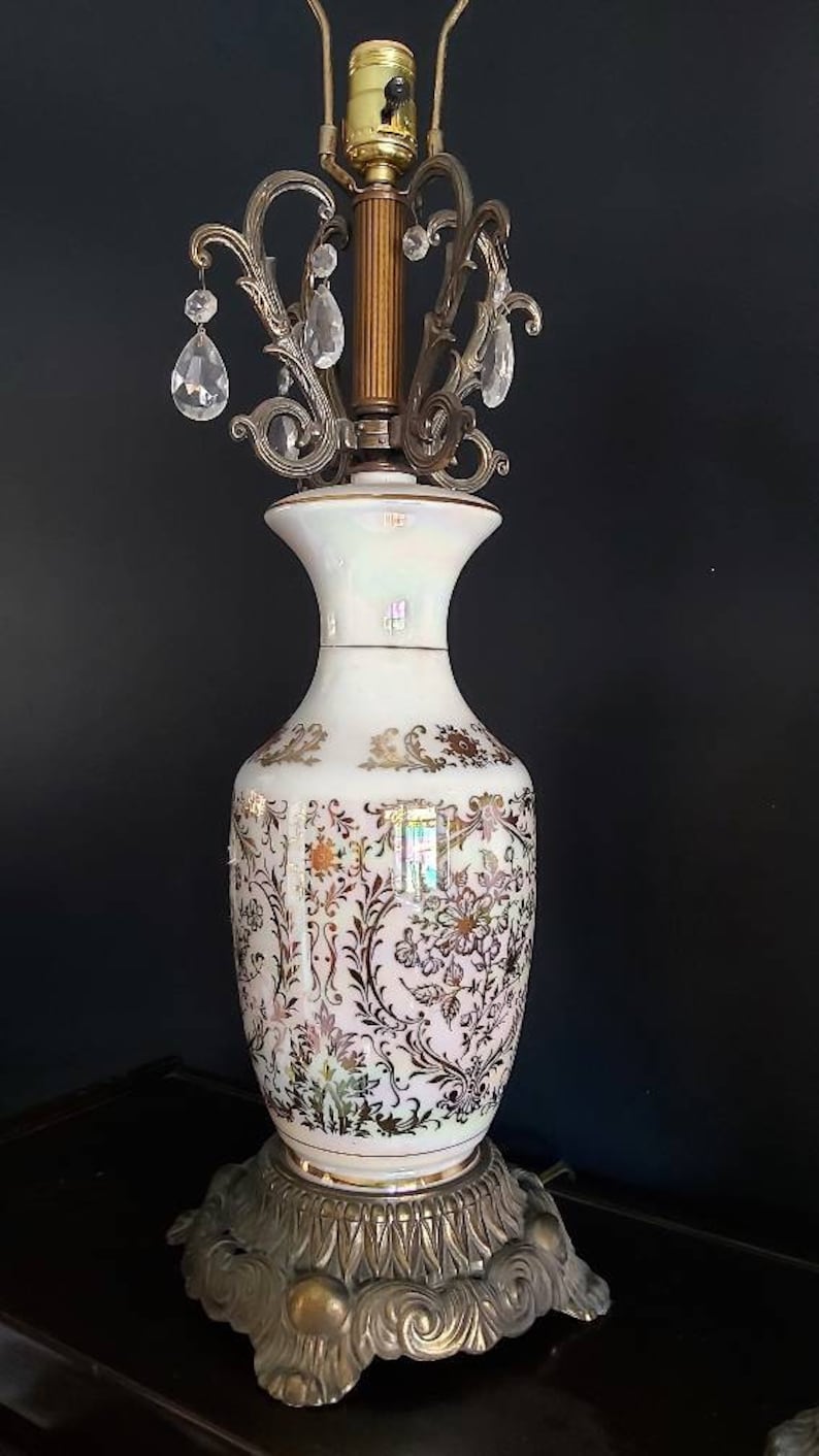 May include: An ornate table lamp with a white ceramic base featuring a floral pattern in gold and brown. The lamp has a brass-colored metal base and a decorative top with hanging crystal accents. The lamp's design is vintage and elegant.